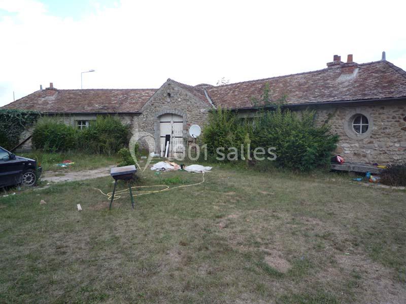 Bâtisse en pierre avec toit en tuiles, entourée de buissons et d'une pelouse, avec des objets épars au sol.