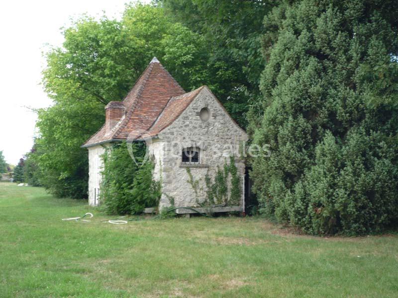 Petite maison en pierre avec toit en tuiles, entourée d'arbres et de végétation, située sur une pelouse.