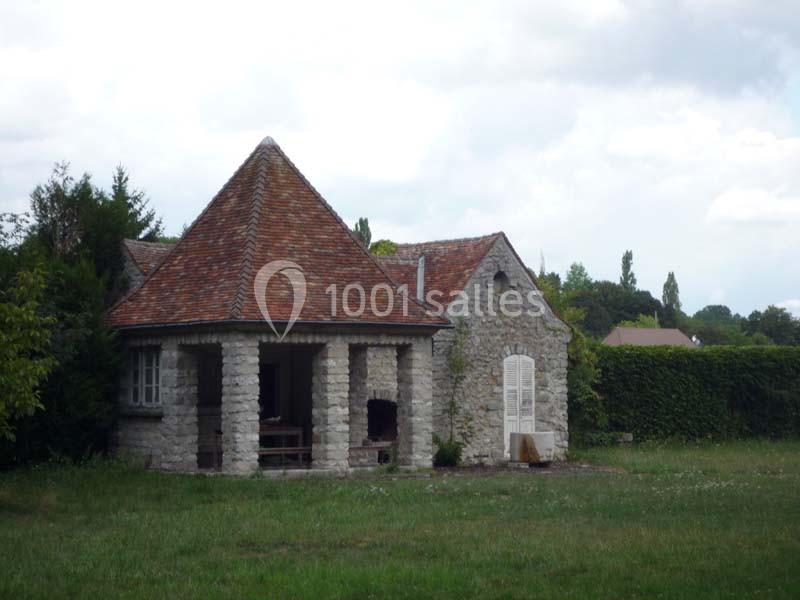 Maison en pierre avec toit en tuiles rouges, porche couvert et jardin verdoyant sous un ciel nuageux.