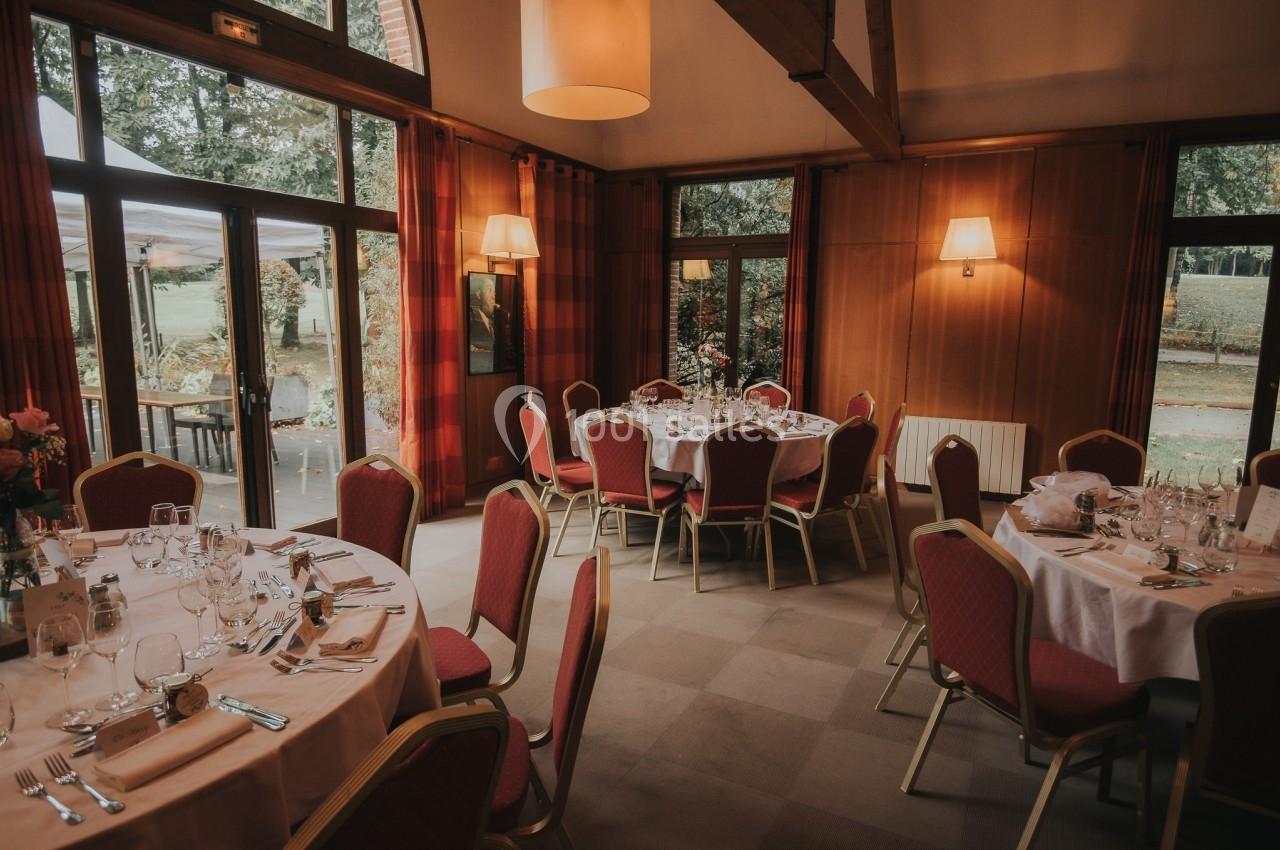 Salle de réception avec tables rondes dressées, chaises rouges et grandes fenêtres donnant sur un jardin.