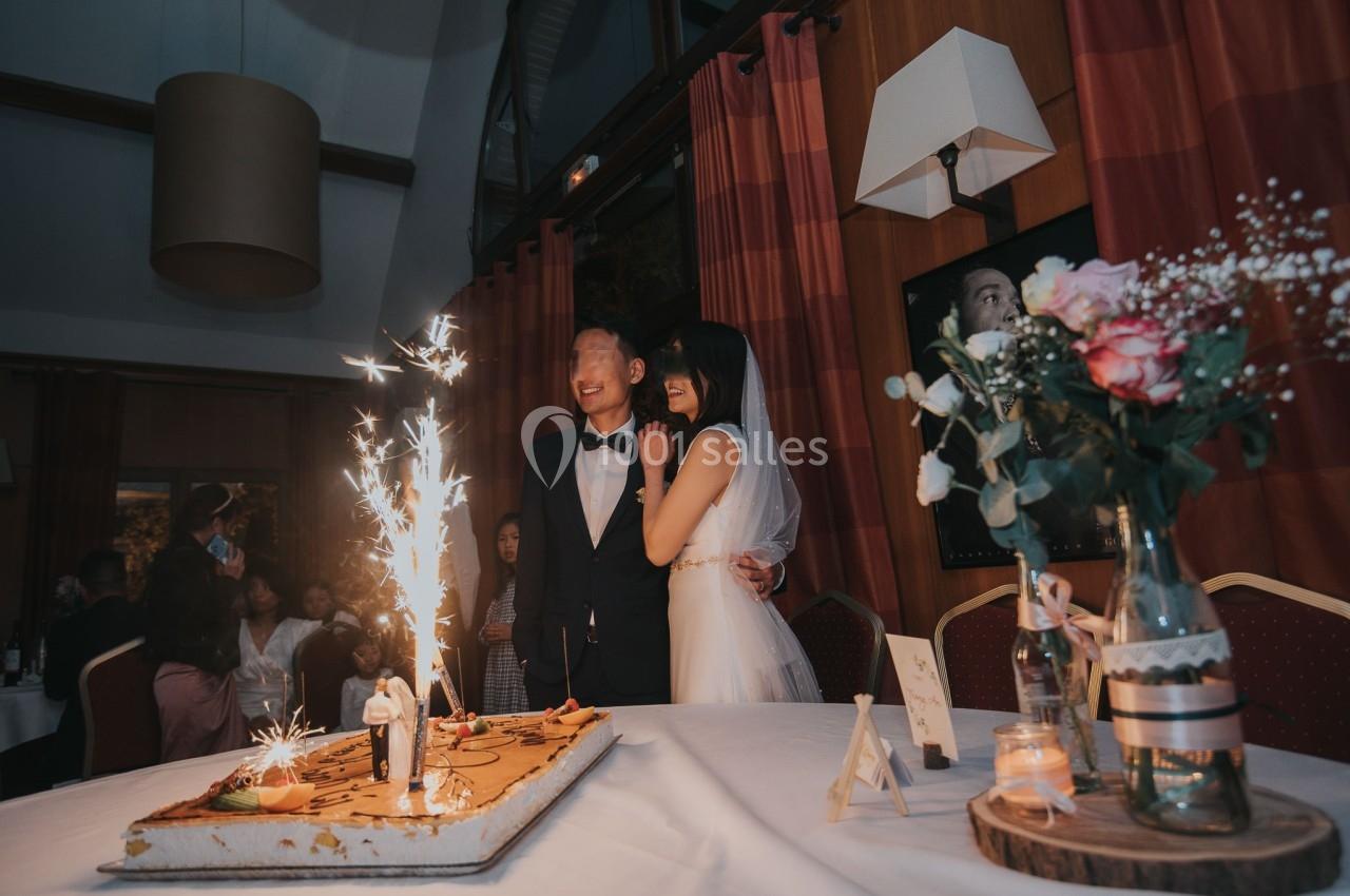 Un couple de mariés souriant devant un gâteau décoré de cierges magiques, entouré d'invités dans une salle de réception.
