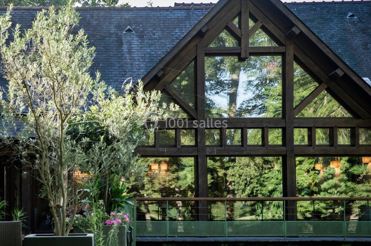 Façade en bois avec de grandes baies vitrées reflétant des arbres, entourée de végétation et d'une terrasse.