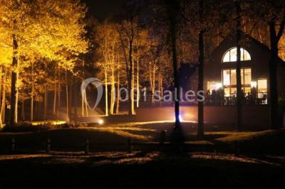Maison en bois éclairée la nuit, entourée d'arbres illuminés, sous un ciel nuageux avec la lune visible.