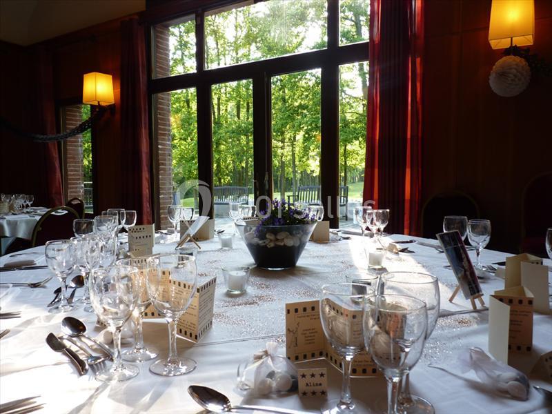 Salle de réception avec tables dressées, nappes blanches, verres, couverts et centre de table floral près de grandes baies…