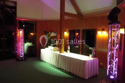 Maison en bois éclairée la nuit, entourée d'arbres illuminés, sous un ciel nuageux avec la lune visible.