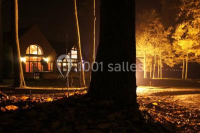 Maison en bois éclairée la nuit, entourée d'arbres illuminés, sous un ciel nuageux avec la lune visible.