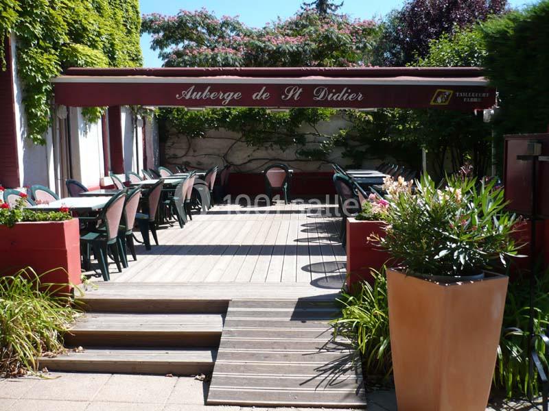 Terrasse extérieure d'un restaurant avec tables et chaises alignées sous un auvent rouge, entourée de plantes.
