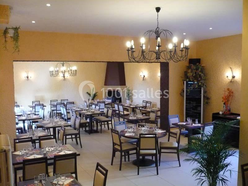 Salle de restaurant lumineuse avec tables dressées, chaises en bois, murs beige, plantes décoratives et lustres en fer forgé.