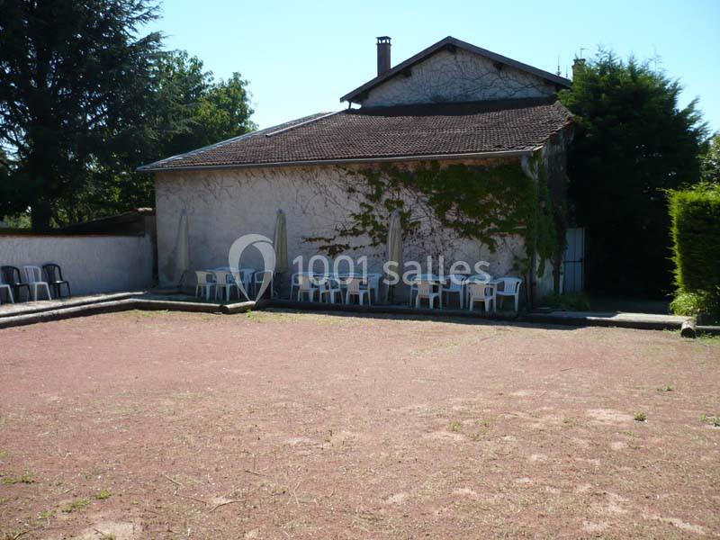 Terrain de pétanque en terre battue devant un bâtiment avec des chaises blanches alignées sous des parasols.