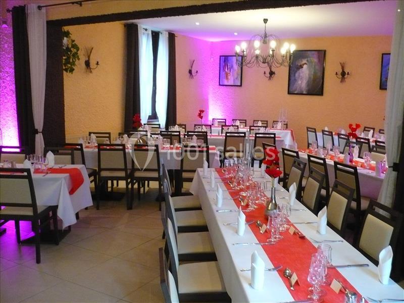 Salle de réception décorée avec des tables dressées, nappes blanches et chemins de table rouges, éclairage chaleureux.
