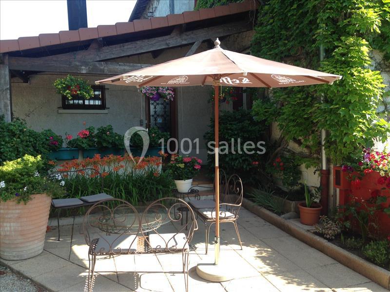 Terrasse avec chaises en métal, parasol beige et plantes fleuries entourant une maison en pierre.