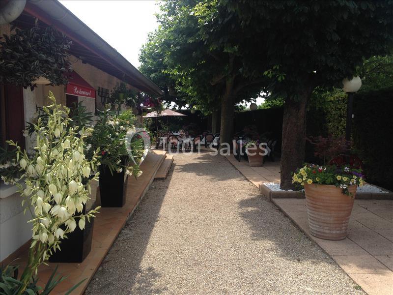 Allée gravillonnée bordée de plantes menant à une terrasse ombragée avec des tables et des chaises.