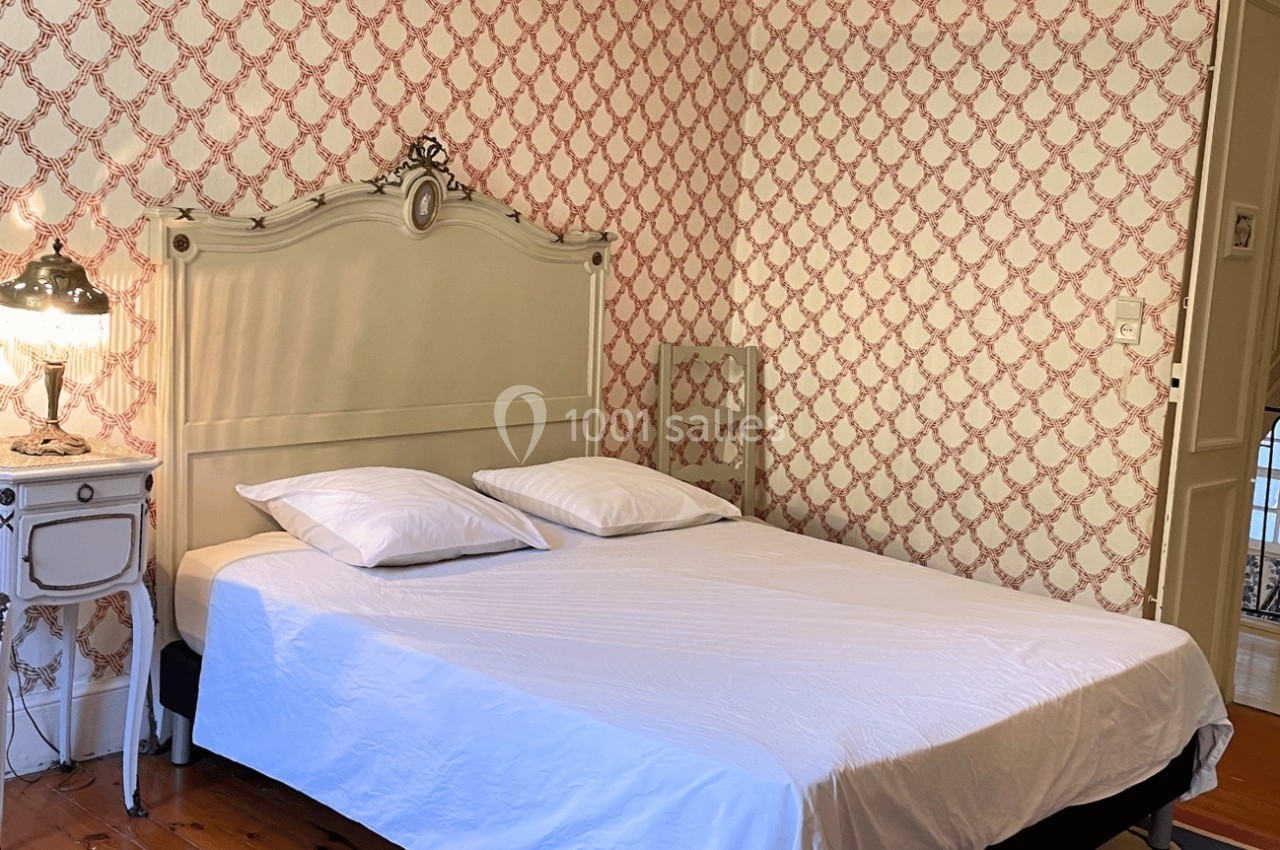 Lit double avec draps blancs dans une chambre au style classique, avec papier peint à motifs rouges et lampe de chevet.