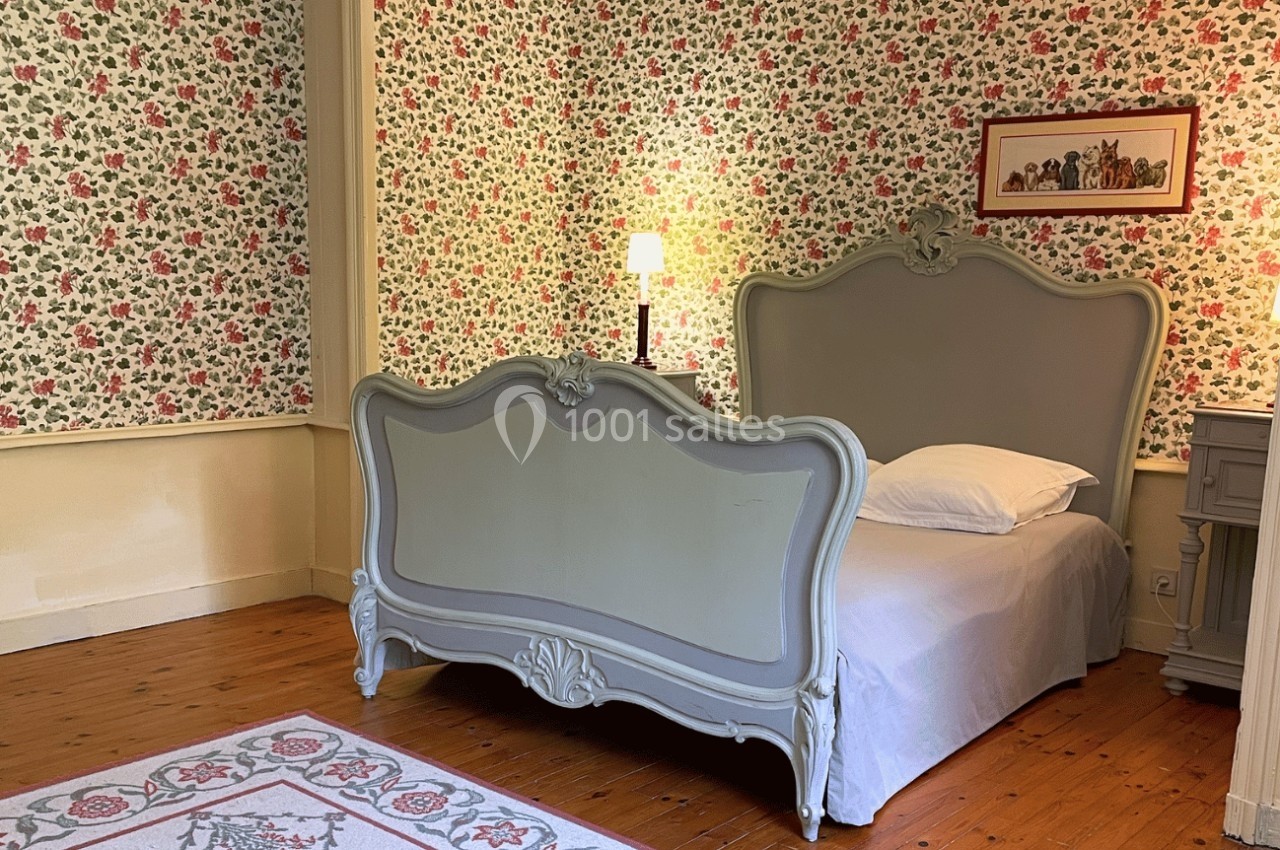 Chambre avec lit ancien gris, mur tapissé de motifs floraux, lampe allumée et tapis décoratif au sol.