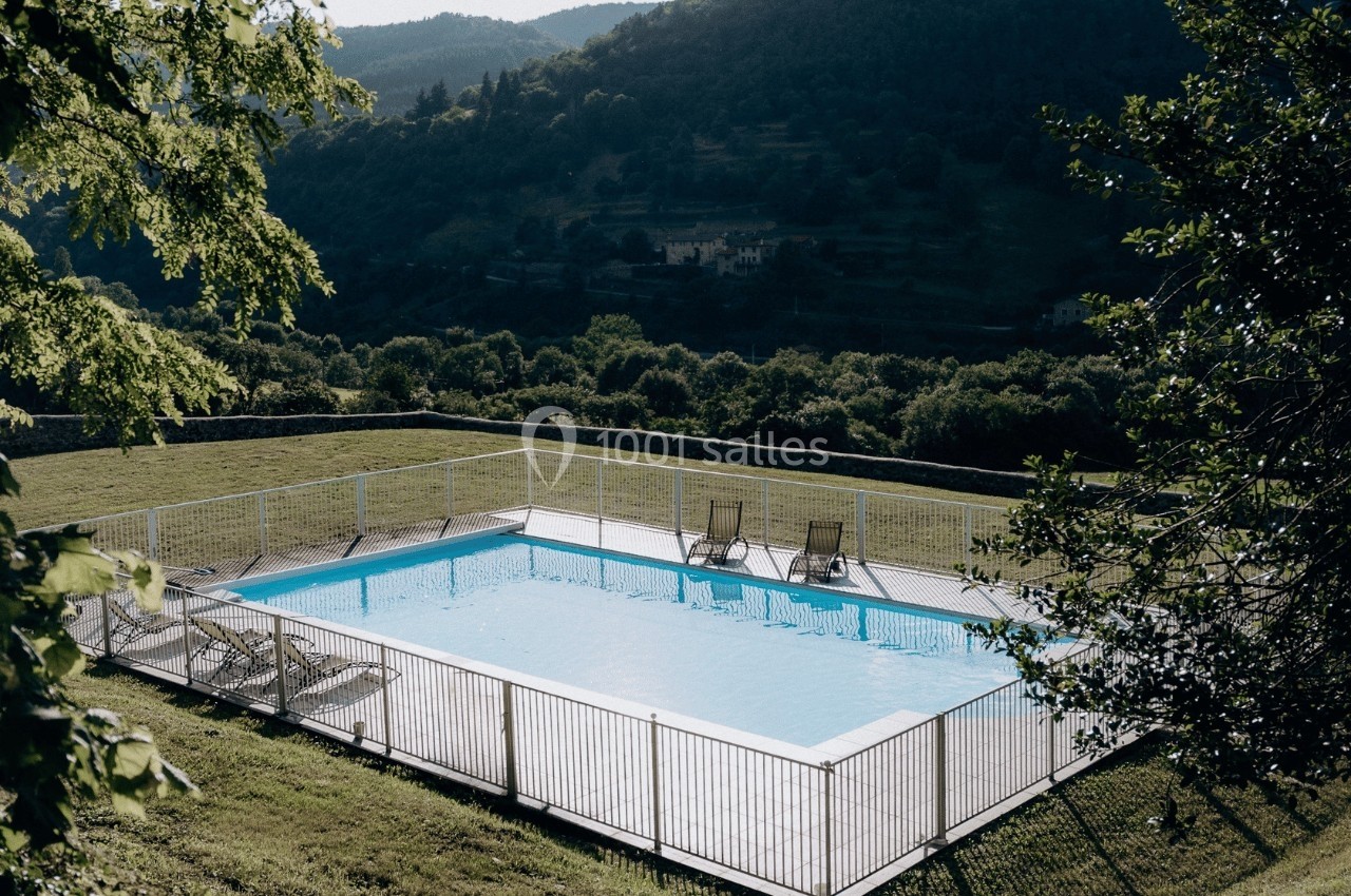 Piscine extérieure entourée d'une clôture, située dans un cadre naturel avec collines verdoyantes en arrière-plan.