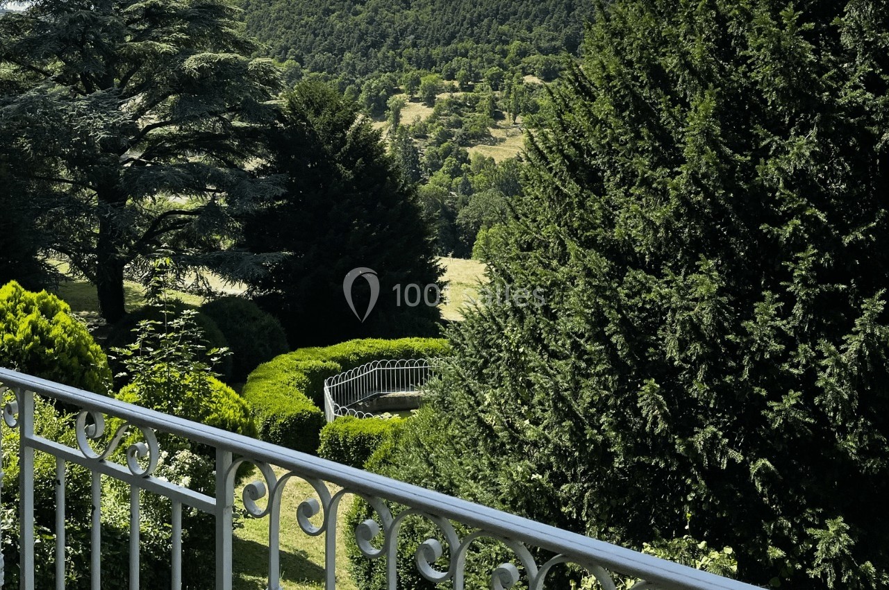 Vue d'un jardin verdoyant avec des arbres, une balustrade blanche au premier plan et des collines en arrière-plan.