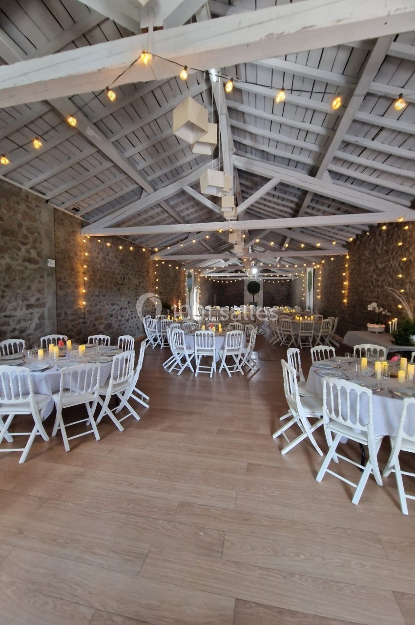 Salle de réception décorée avec des tables rondes, nappes blanches, chaises blanches et guirlandes lumineuses suspendues.