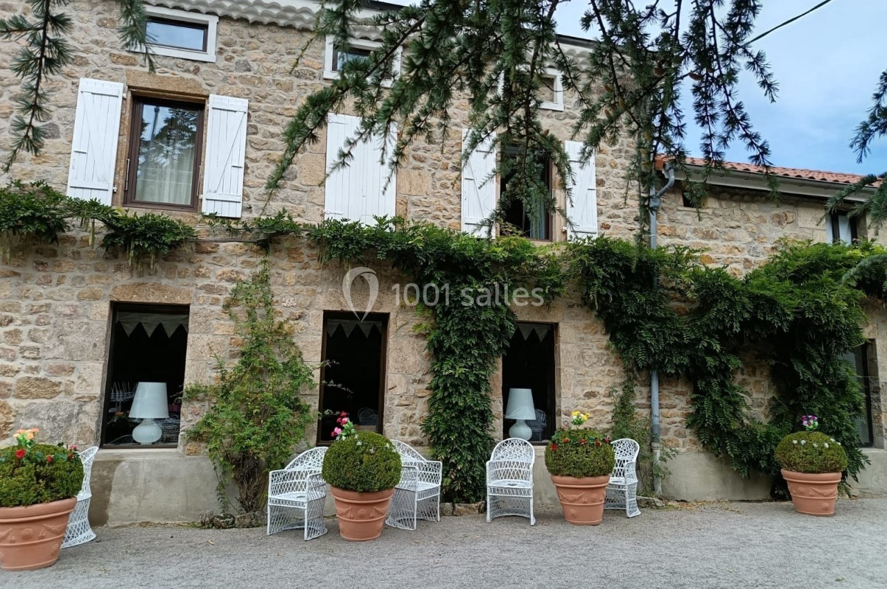 Façade en pierre d'une maison avec volets blancs, plantes grimpantes et chaises blanches disposées devant.