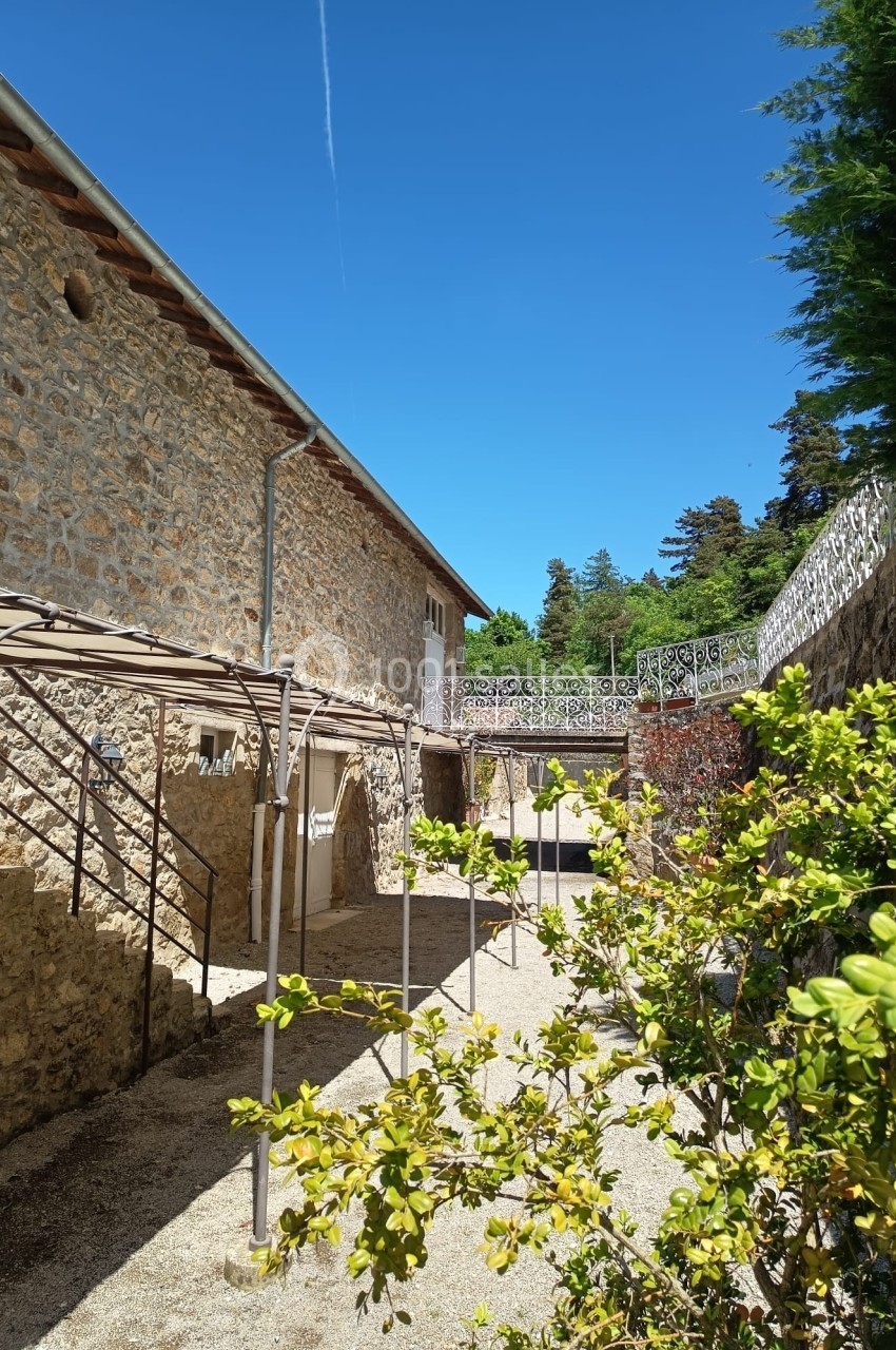 Cour extérieure ensoleillée avec murs en pierre, pergola métallique, arbustes et ciel bleu dégagé.