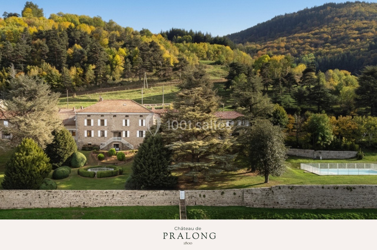 Vue d'un château en pierre entouré de jardins, d'arbres et de collines boisées sous un ciel dégagé.
