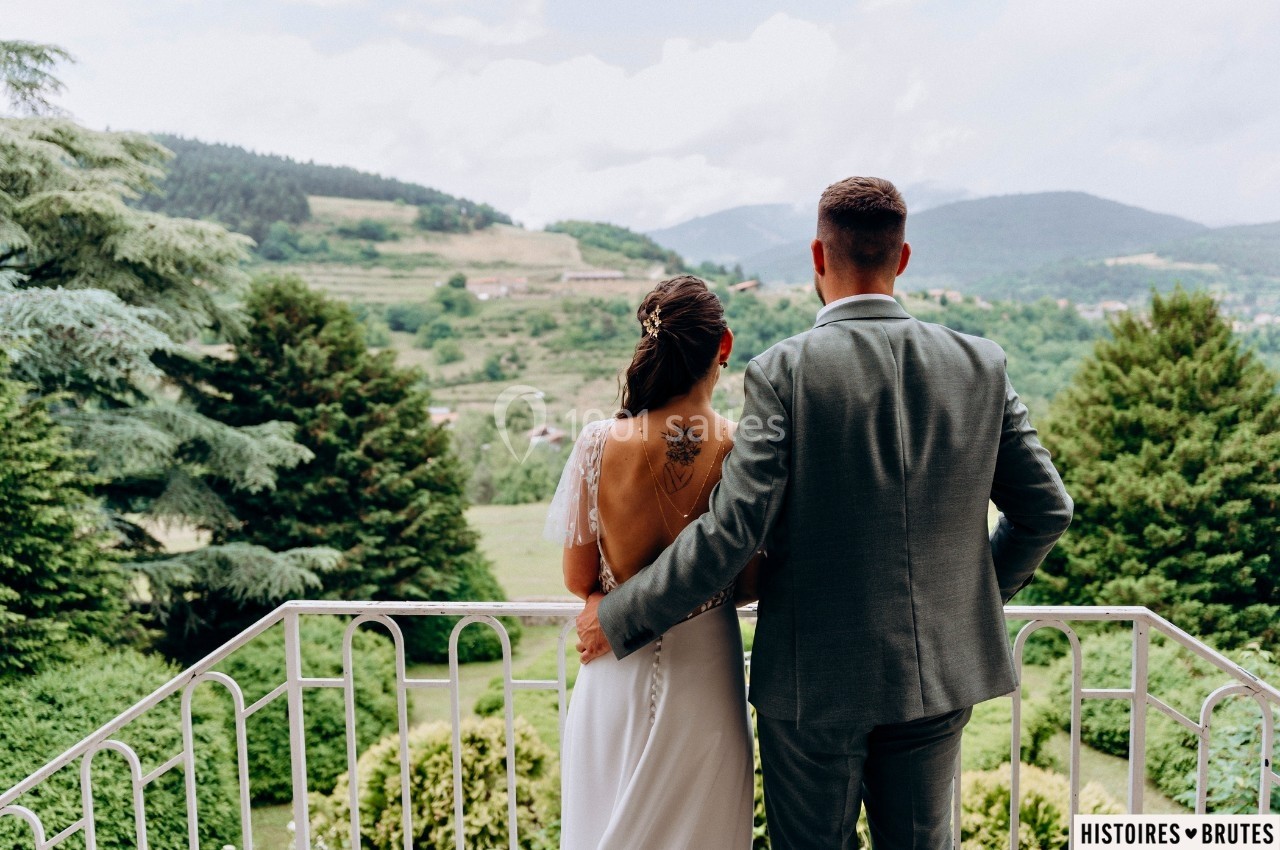 Un couple de dos, debout sur un balcon, regarde un paysage vallonné avec des arbres et des collines.