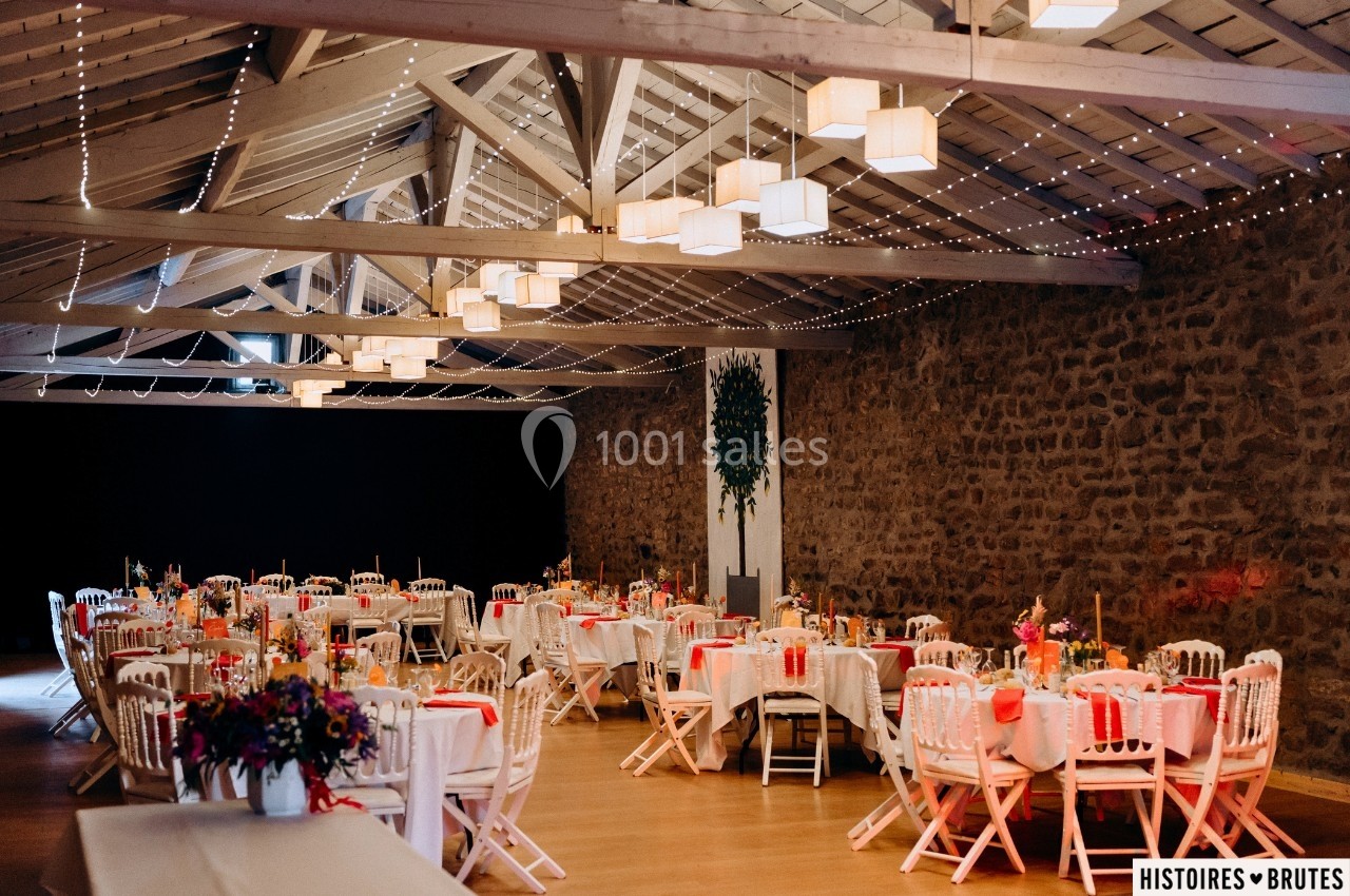 Salle de réception décorée pour un événement, avec tables dressées, chaises blanches et guirlandes lumineuses suspendues.