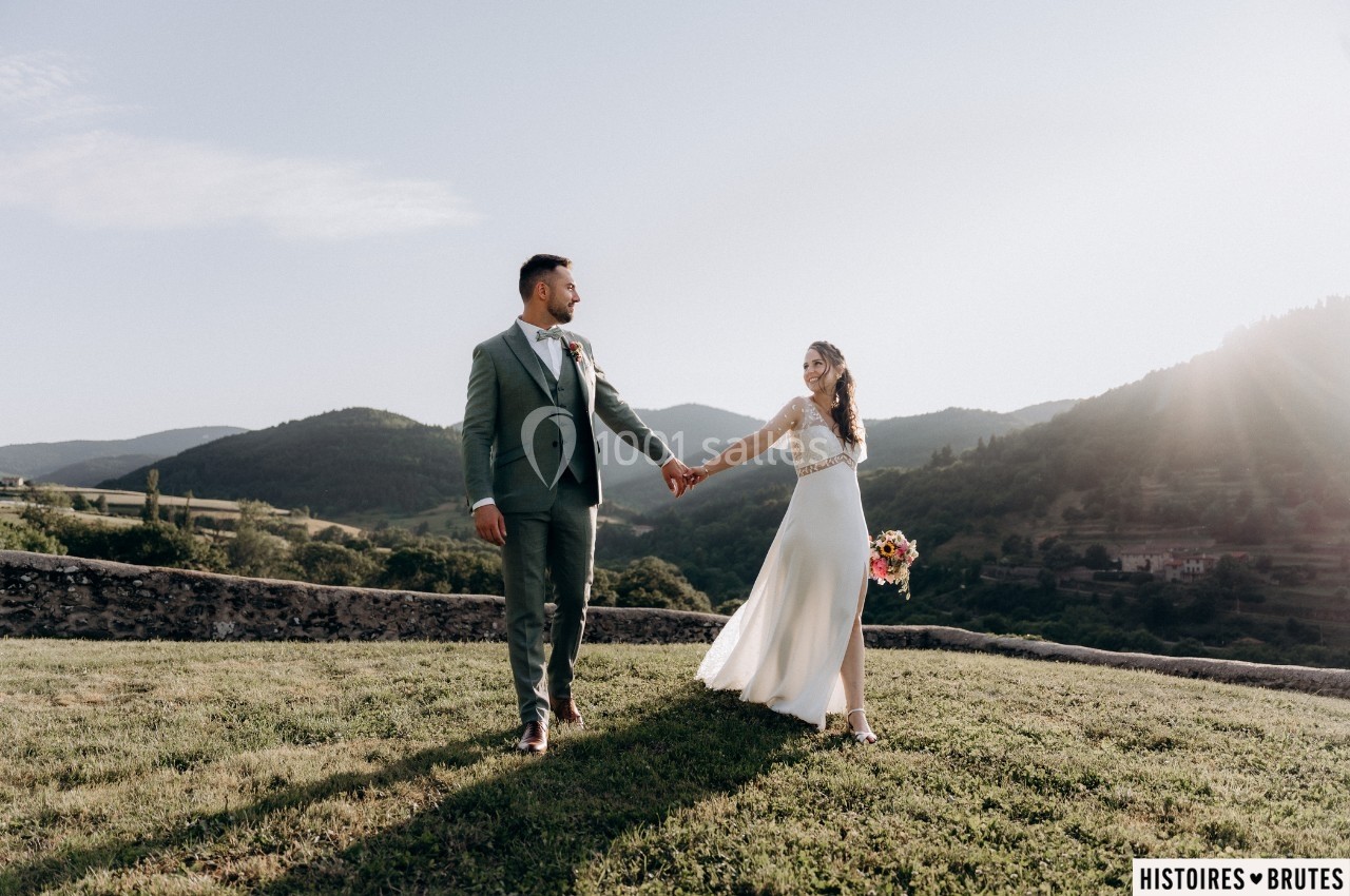 Un couple en tenue de mariage marche main dans la main dans un paysage vallonné sous un ciel dégagé.