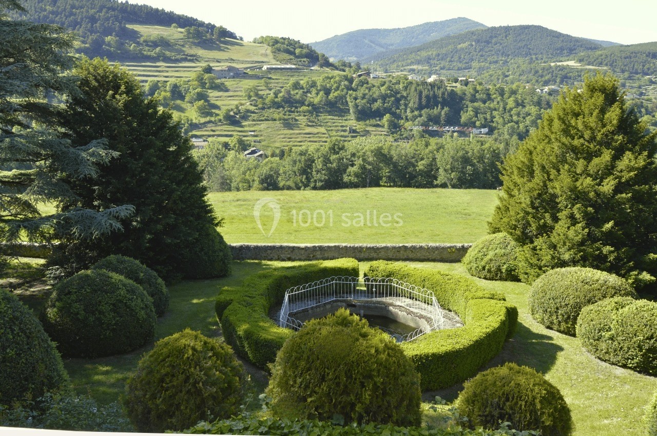 Vue d'un jardin paysager avec une fontaine centrale entourée de haies, et des collines verdoyantes en arrière-plan.