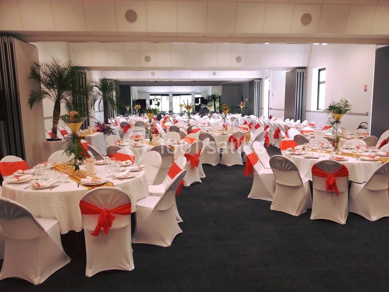 Salle de réception décorée avec des tables rondes dressées, nappes blanches et chaises ornées de nœuds orange.