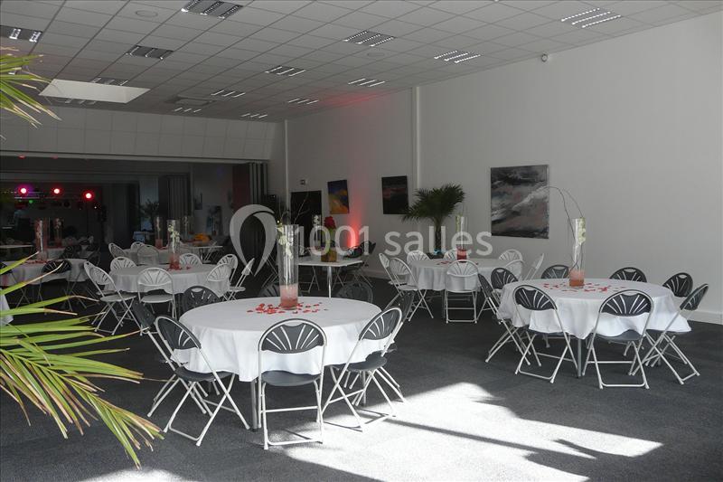 Salle de réception lumineuse avec tables rondes dressées, nappes blanches et décorations simples.