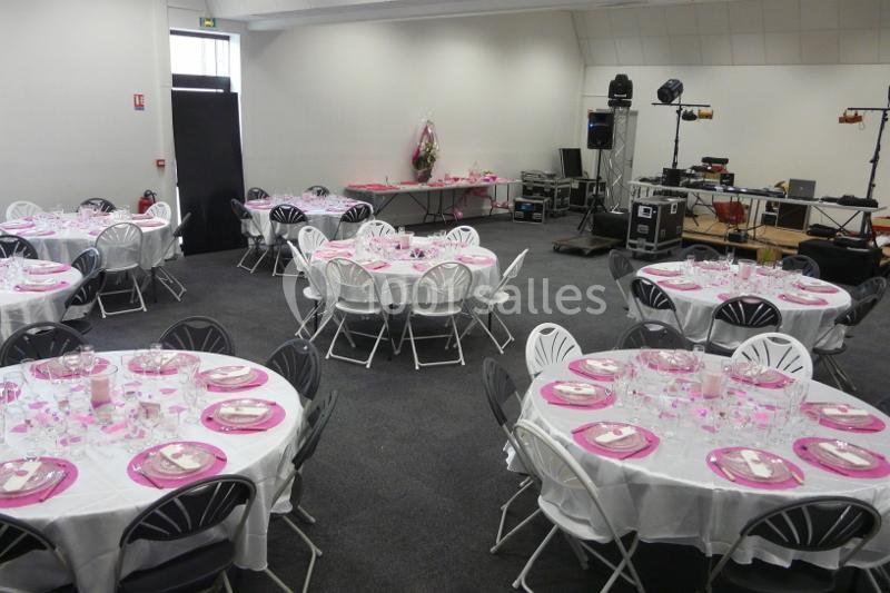 Salle décorée pour un événement avec des tables rondes dressées de nappes blanches et d'assiettes roses.