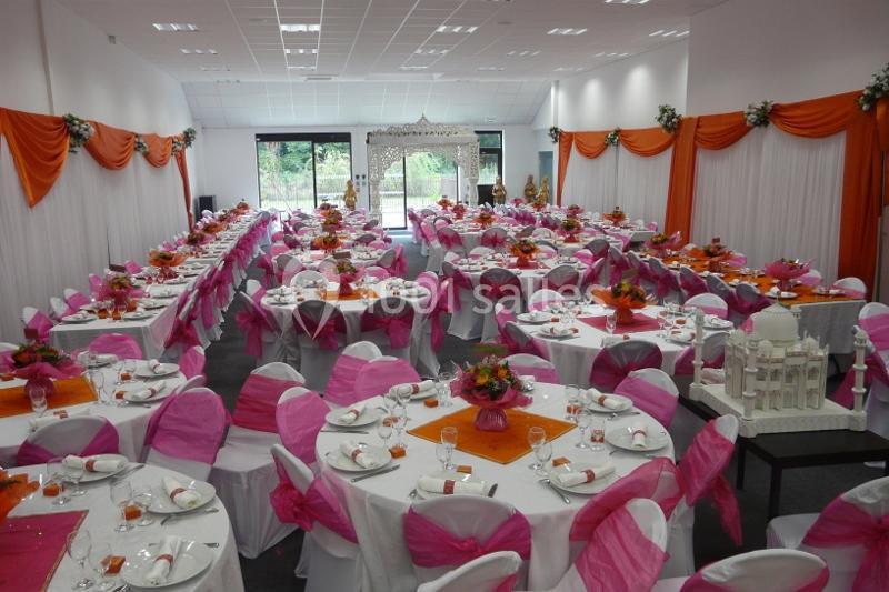 Salle décorée pour un événement, avec des tables rondes dressées, nappes blanches, chaises roses et orange, et compositions…