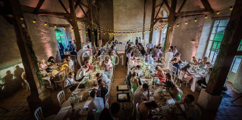 Salle de réception rustique avec de longues tables décorées, occupées par des invités lors d'un événement festif.