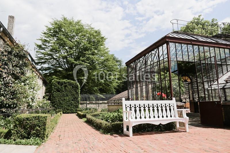 Allée pavée bordée de haies, avec une serre vitrée et un banc blanc dans un jardin arboré.