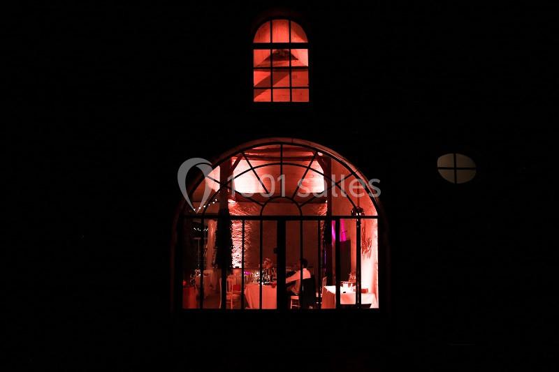 Vue nocturne d'une salle éclairée en rouge à travers une grande baie vitrée cintrée.