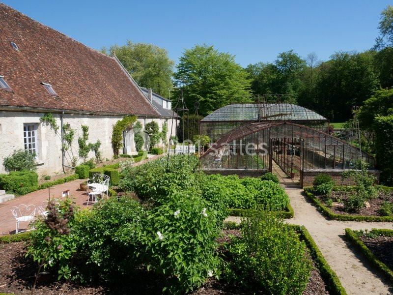 Jardin avec allées bordées de plantes, serre en verre et bâtiment en pierre sous un ciel dégagé.