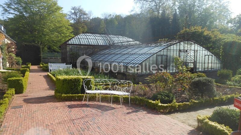Serres vitrées entourées de haies taillées et d'un chemin pavé, avec un banc en fer forgé au premier plan.