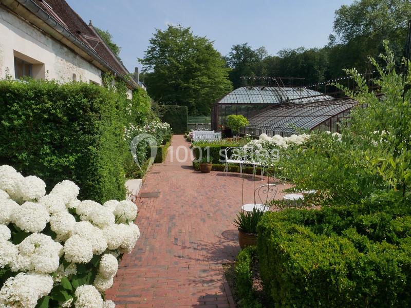 Allée en briques bordée de buissons fleuris, menant à des serres vitrées entourées de verdure.