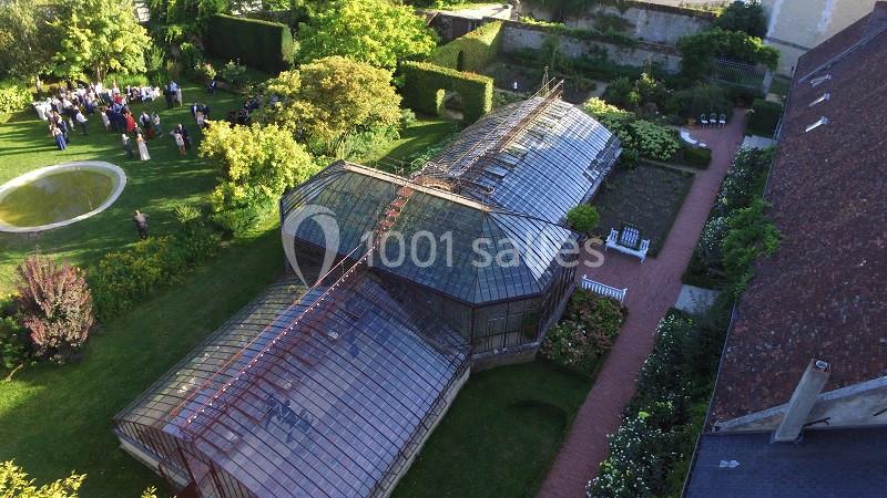 Vue aérienne d'une serre en verre entourée de jardins verdoyants avec des allées et un groupe de personnes au loin.