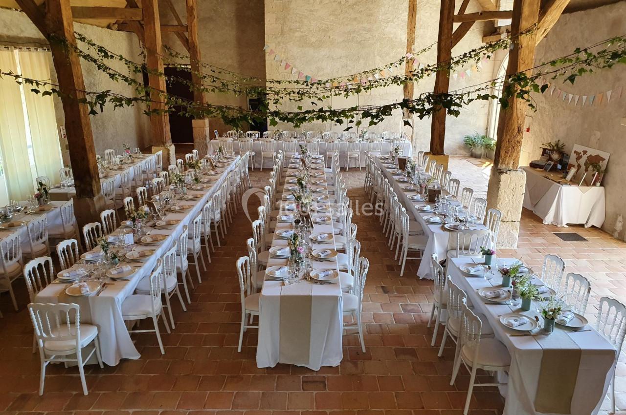 Salle de réception décorée pour un mariage, avec de longues tables dressées, nappes blanches et guirlandes suspendues.