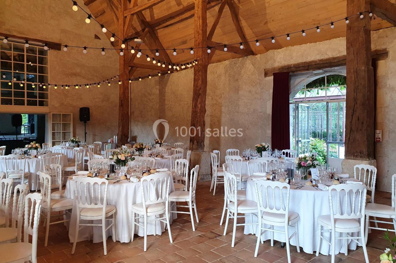 Salle de réception rustique avec tables rondes dressées, chaises blanches et guirlandes lumineuses suspendues.