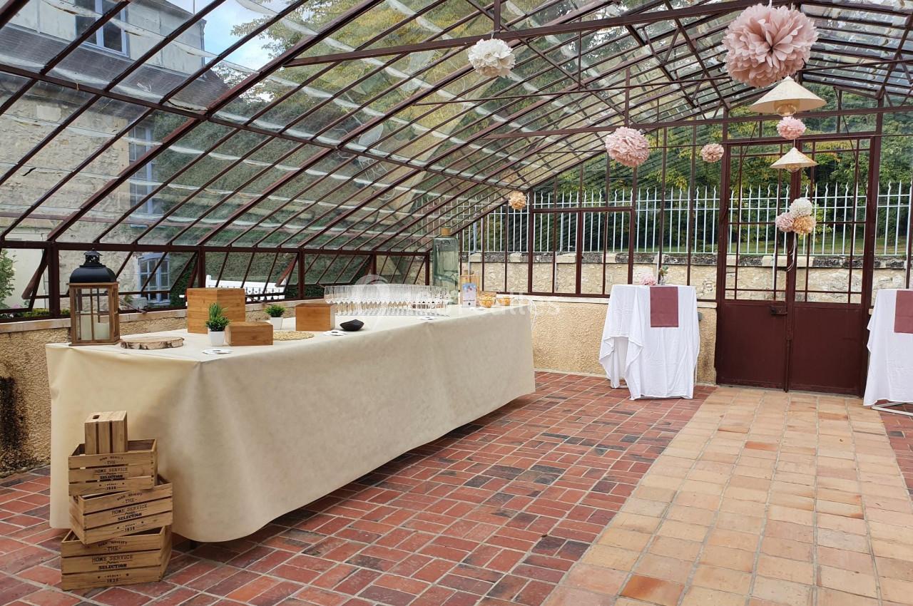 Salle en verrière aménagée pour un événement, avec table de buffet, décorations suspendues et sol en briques.