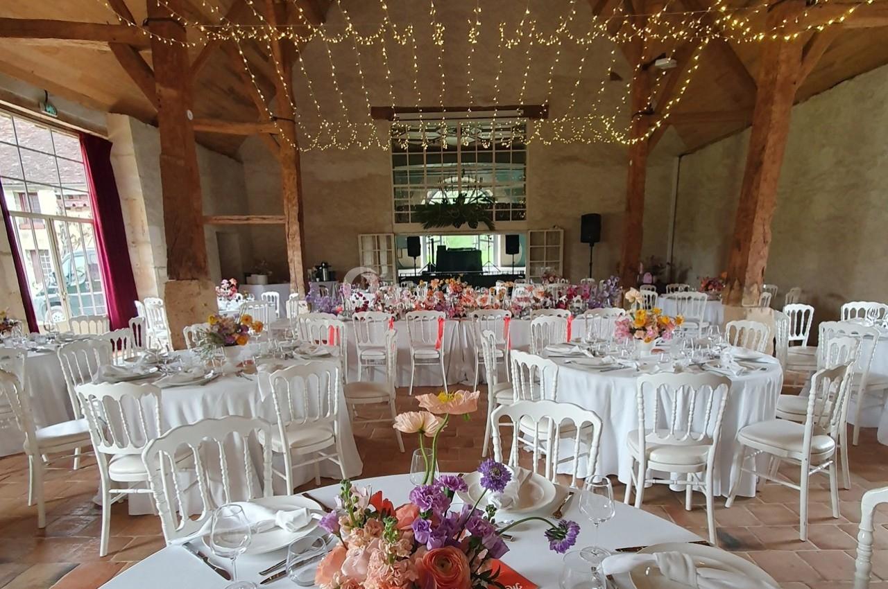 Salle de réception décorée pour un mariage, avec tables rondes ornées de fleurs et guirlandes lumineuses suspendues.