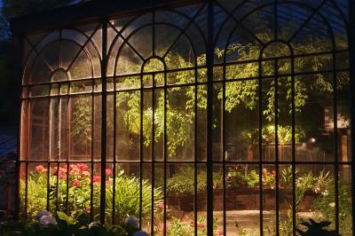 Serre illuminée au crépuscule, entourée de murs en pierre et d'arbres, avec une tour en arrière-plan.