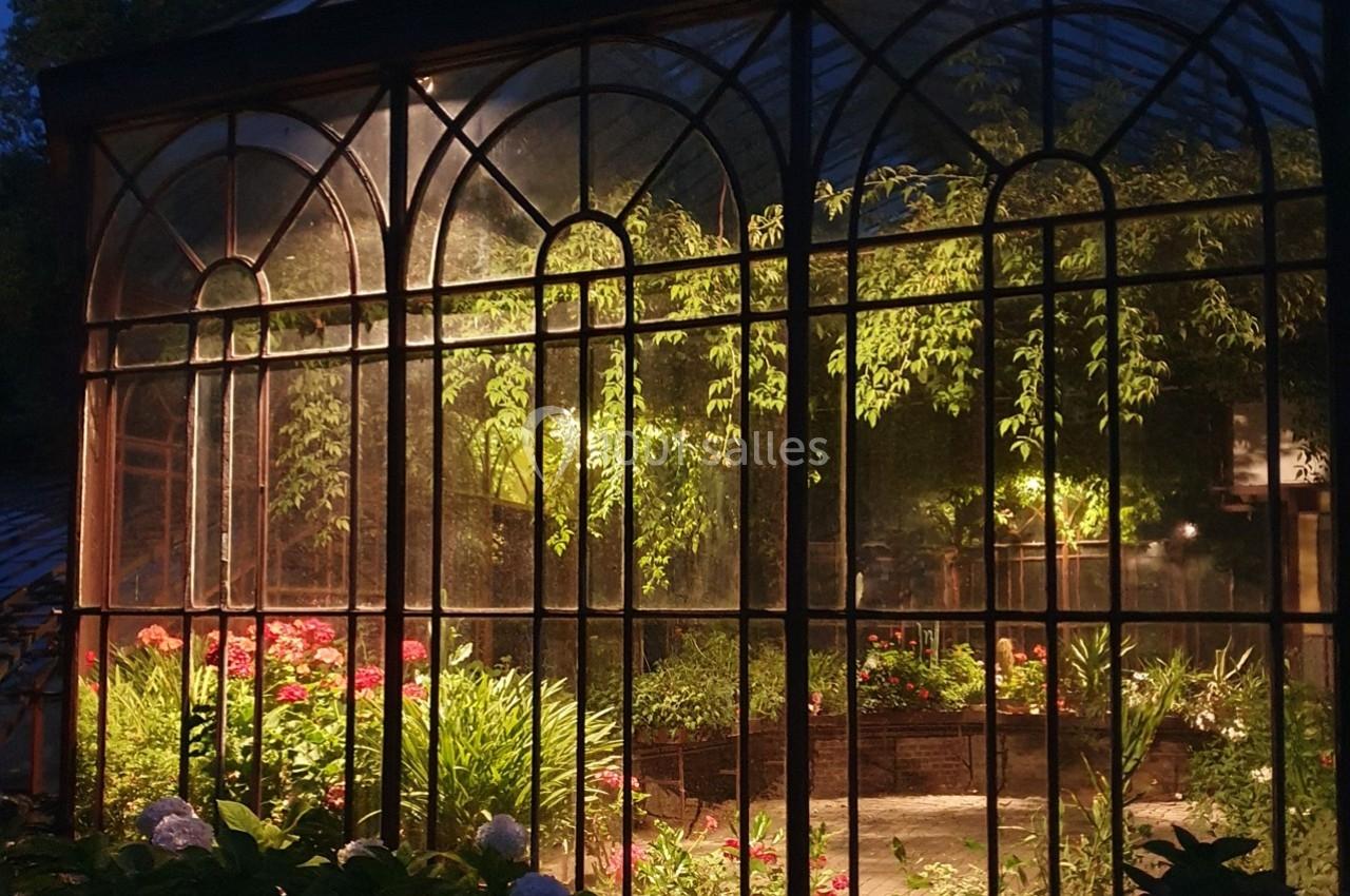 Serre éclairée de nuit avec des plantes verdoyantes et des fleurs visibles à travers des vitres encadrées de métal.