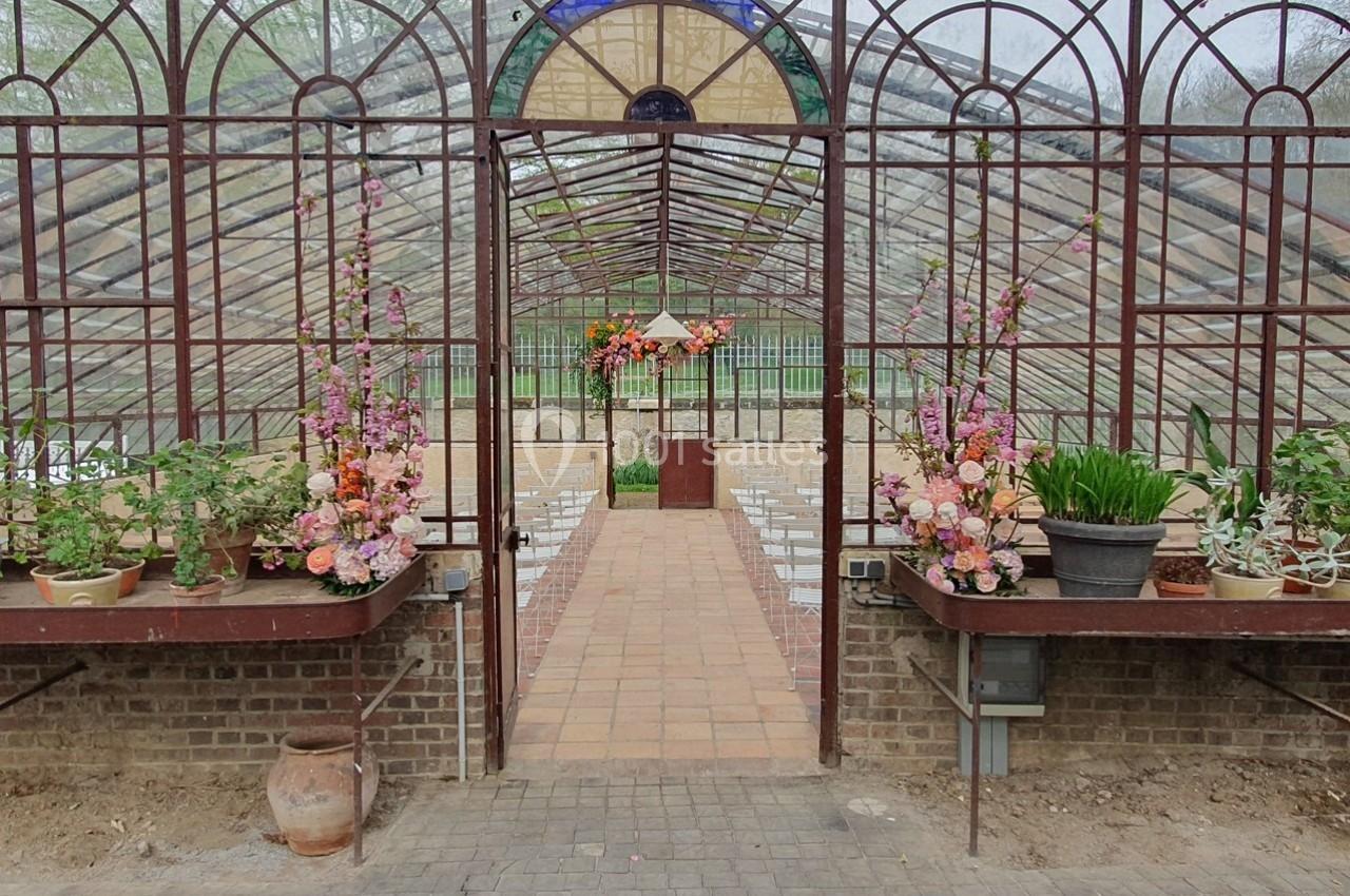 Serre avec structure métallique, allée centrale en briques, fleurs décoratives et plantes en pots sur des étagères.