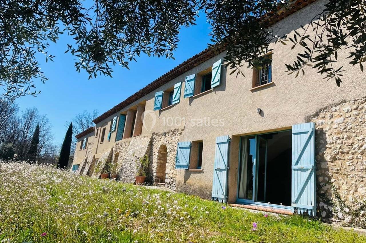 Façade d'une maison en pierre avec volets bleus, entourée d'un champ fleuri sous un ciel ensoleillé.