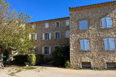 Bâtisse en pierre avec toit en tuiles, entourée de végétation et d'un chemin sous un ciel bleu clair.