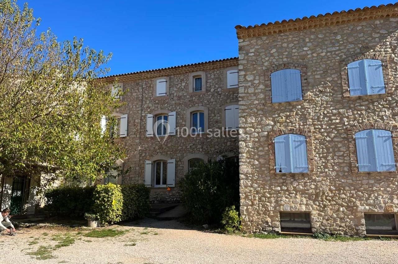 Bâtiment en pierre avec volets bleus, entouré d'arbres et donnant sur une cour ensoleillée.