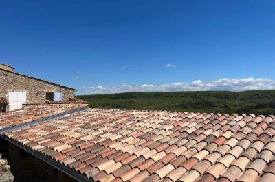 Bâtisse en pierre avec toit en tuiles, entourée de végétation et d'un chemin sous un ciel bleu clair.
